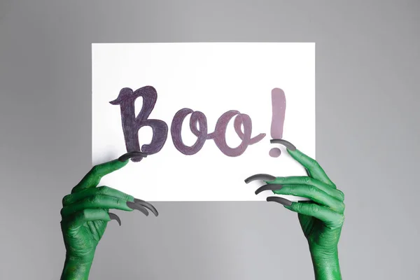 Ruce čarodějnice držící papír s textem Boo na šedém pozadí. Halloween oslava — Stock fotografie