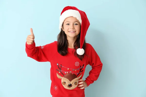 Malá holka ve vánočním svetru a Santa klobouk ukazující palec nahoru na barevném pozadí — Stock fotografie