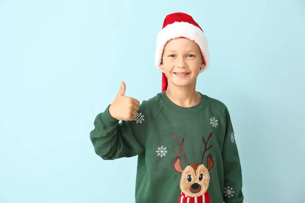 Menino em camisola de Natal e chapéu de Papai Noel mostrando polegar para cima no fundo de cor — Fotografia de Stock