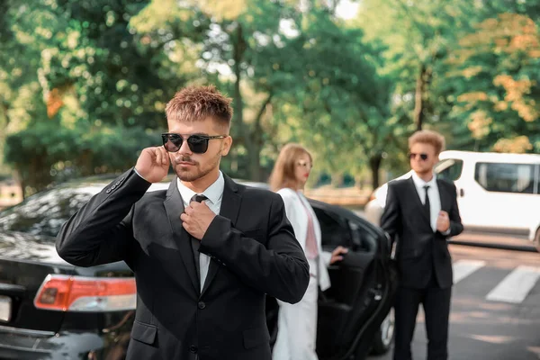 Young celebrity with bodyguards near car outdoors — Stock Photo, Image
