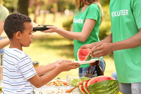 Young volunteer giving food to African-American poor little boy outdoors