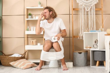 Young man pinching nose while sitting on toilet bowl at home clipart