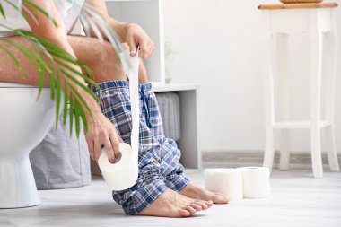 Evdeki klozetin üzerinde kağıt rulo oturan genç adam.