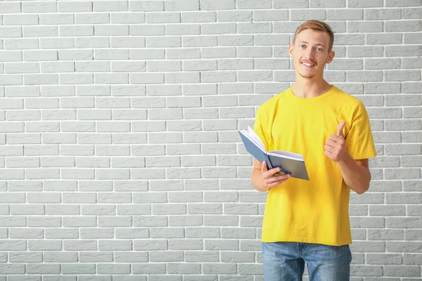 Młody mężczyzna w stylowej koszulce i z książką pokazującą gest kciuka w górę na ceglanym tle — Zdjęcie stockowe