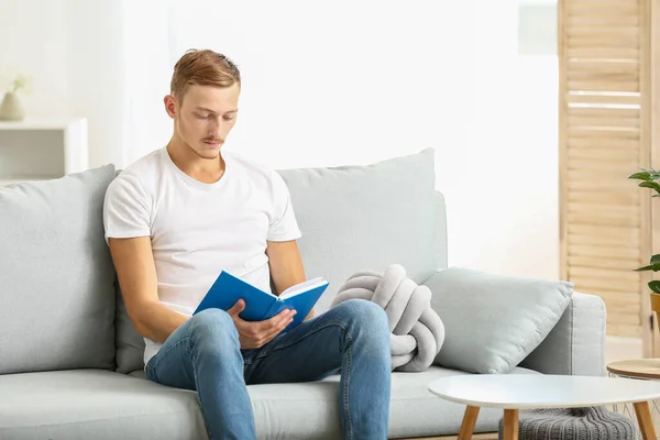 Young man in stylish t-shirt and with book sitting on sofa at home — Stock Photo, Image