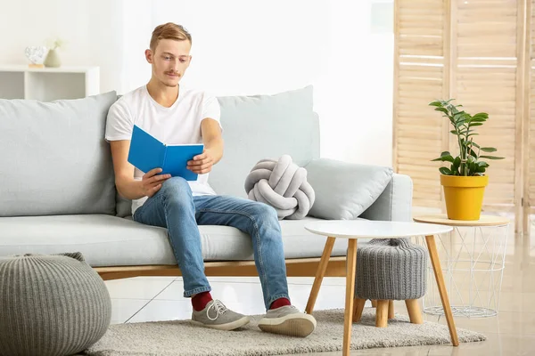 Young man in stylish t-shirt and with book sitting on sofa at home — Stock Photo, Image