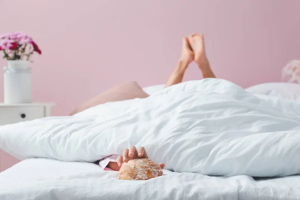 Young woman reaching hand for tasty bun while lying in bed — Stock Photo, Image