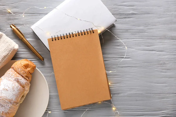 Gri ahşap arka planda kalem, kruvasan ve çelenk bulunan defterler.