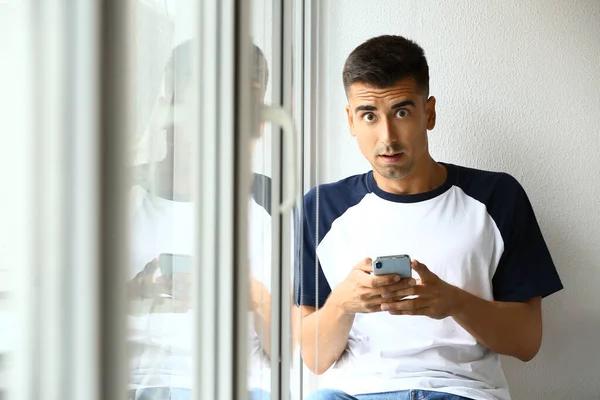 Surprised young man with mobile phone near window