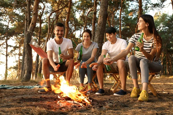 Young friends roasting sausages on fire and drinking beer at camp — Stock Photo, Image