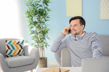 Evde bilgisayarla çalışırken telefonla konuşan yakışıklı bir adam.