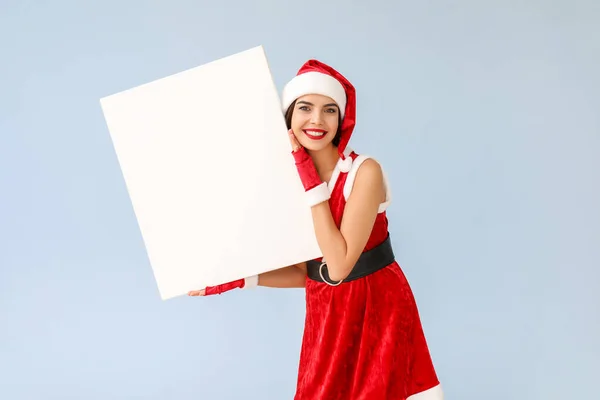 Beautiful young woman in Santa Claus costume and with blank poster on light background — Stock Photo, Image