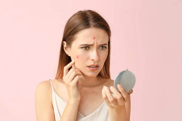 Portrait of young woman with acne problem looking in mirror on color background — Stock Photo, Image