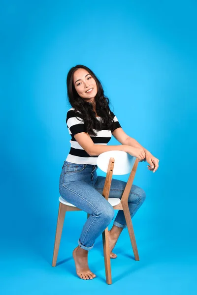 Beautiful Asian woman sitting on chair against color background — Stock Photo, Image