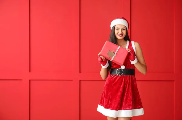 Beautiful young woman in Santa Claus costume and with gift near color wall — Stock Photo, Image