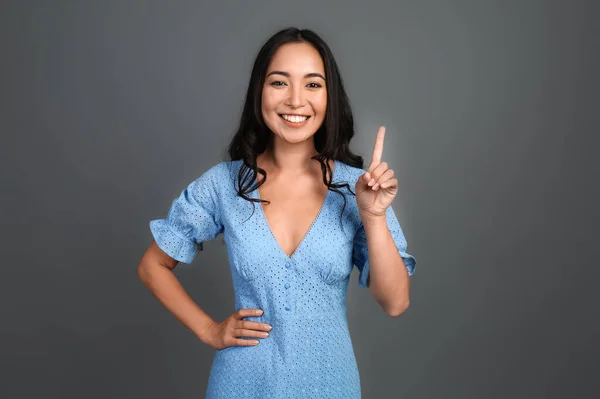 Beautiful Asian woman with raised index finger on grey background — Stock Photo, Image