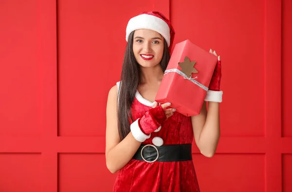 Hermosa mujer joven en traje de Santa Claus y con regalo cerca de la pared de color —  Fotos de Stock