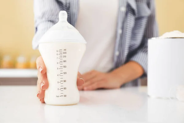 Mother with bottle of baby milk formula on table in kitchen, closeup — Stock Photo, Image