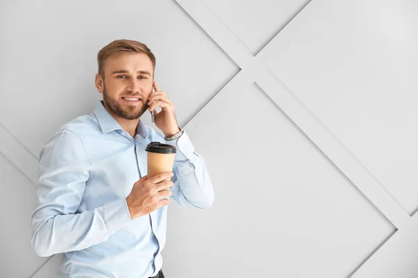 Portrait of handsome businessman with cup of coffee talking by phone on light background — Stock Photo, Image