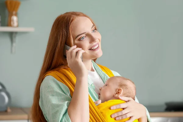 Young mother with little baby in sling talking by mobile phone at home — Stock Photo, Image
