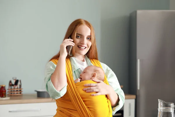 Giovane madre con piccolo bambino in fionda che parla dal telefono cellulare a casa — Foto Stock