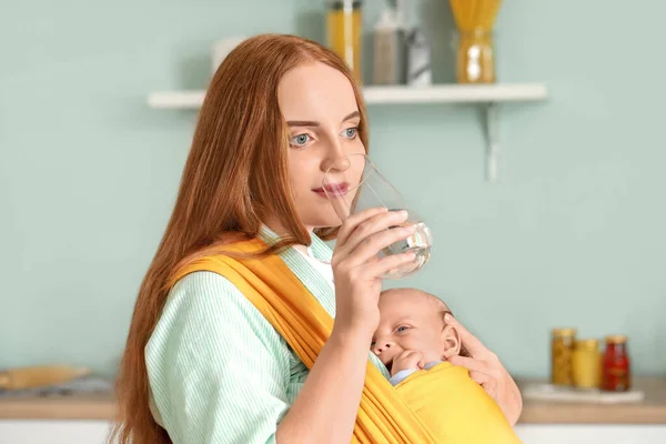 Young mother with little baby in sling drinking water at home — Stock Photo, Image