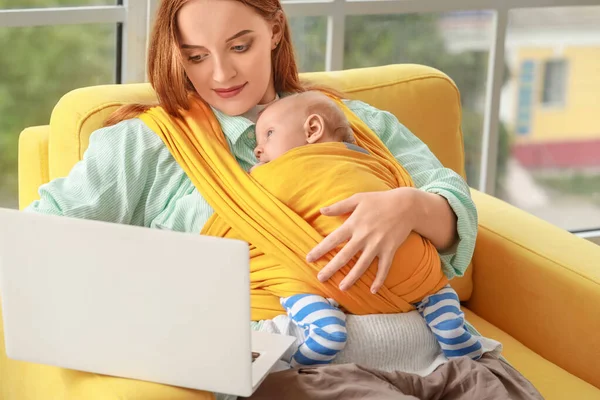 Mamă tânără cu bebeluș în sling folosind laptopul acasă — Fotografie, imagine de stoc