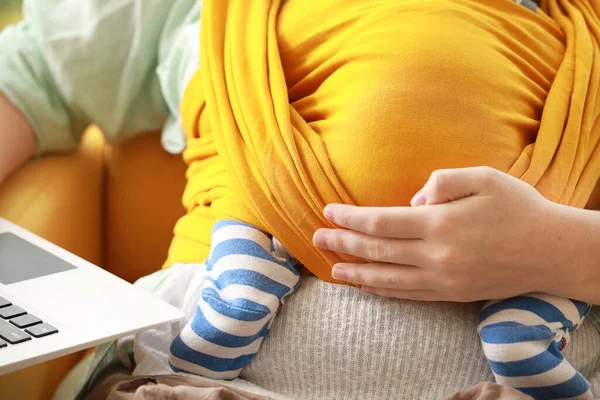 Young mother with little baby in sling using laptop at home, closeup — Stock Photo, Image