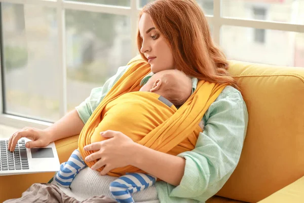Joven madre con un pequeño bebé en cabestrillo usando el portátil en casa — Foto de Stock