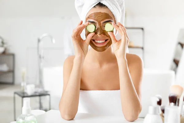 Beautiful young woman with facial mask and cucumber slices in bathroom — Stock Photo, Image