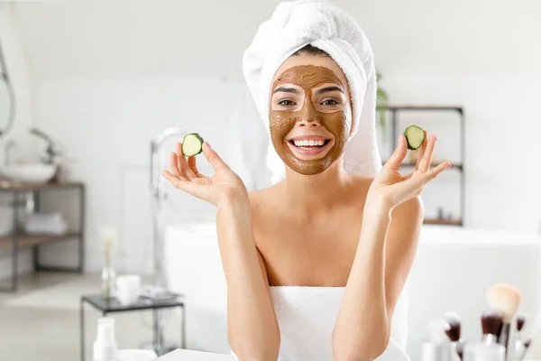 Wanita muda cantik dengan masker wajah dan irisan mentimun di kamar mandi — Stok Foto