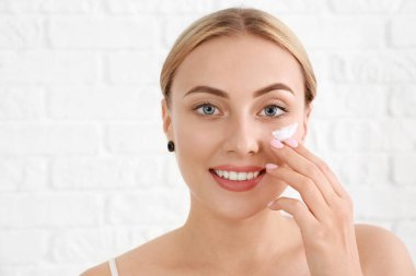 Beautiful young woman applying facial cream against white background