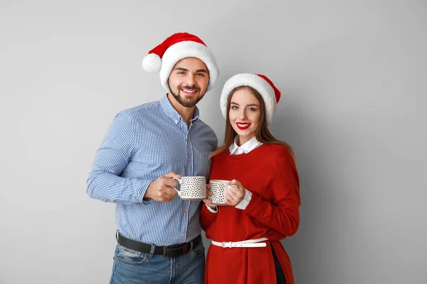 Jovem casal feliz em chapéus de Santa e com chocolate quente no fundo claro — Fotografia de Stock