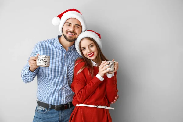 Jovem casal feliz em chapéus de Santa e com chocolate quente no fundo claro — Fotografia de Stock