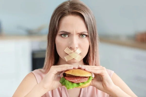 Sad woman with taped mouth and with tasty burger in kitchen. Diet concept