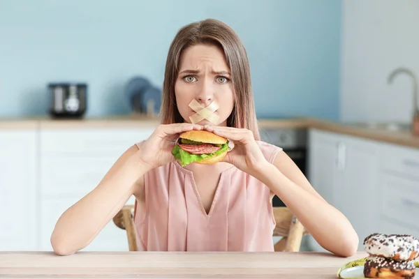 Triste femme à la bouche collée et au délicieux hamburger dans la cuisine. Concept de régime alimentaire — Photo