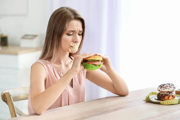 입에 테이프를 붙이고 맛있는 버거를 주방에 갖고 있는 슬픈 여자. 식생활의 개념 — 스톡 사진
