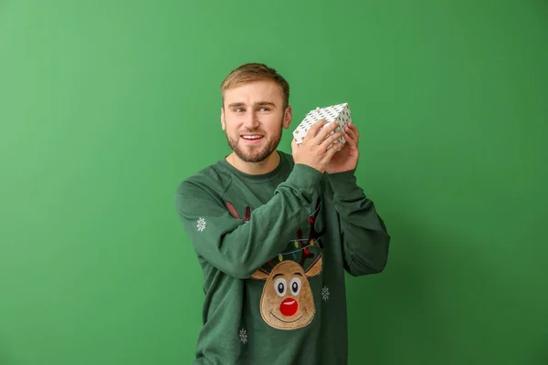 Young man in Christmas sweater and with gift on color background — Stock Photo, Image