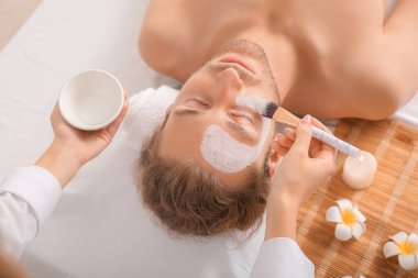 Kozmetik uzmanı spa salonundaki adamın yüzüne maske takıyor.