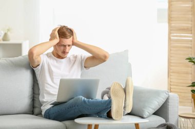 Stresli, şık tişörtlü ve dizüstü bilgisayarlı bir adam evdeki kanepede oturuyor.
