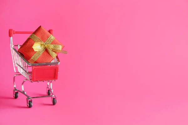Carrinho de compras pequeno com caixa de presente sobre fundo de cor — Fotografia de Stock