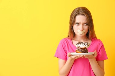 Ağzı bantlanmış üzgün bir kadın ve arka planda lezzetli çörekler. Diyet kavramı