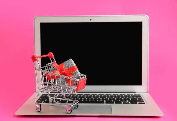 Laptop and small cart with gift on color background. Internet shopping concept — Stock Photo, Image