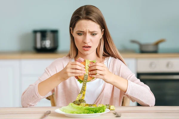 Mujer estresada sosteniendo hamburguesa malsana con cinta métrica en la cocina. Concepto de dieta — Foto de Stock