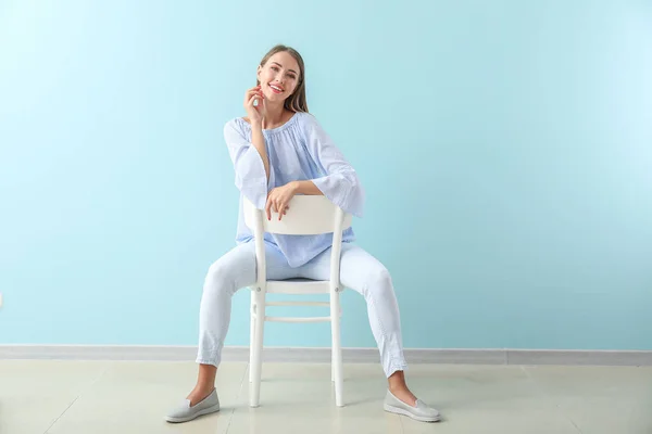 Beautiful young woman sitting on chair against color wall — Stock Photo, Image