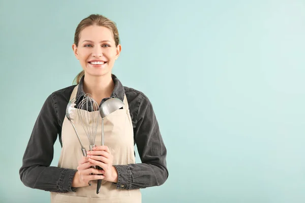 Beautiful young woman in apron and with kitchenware on color background — Stock Photo, Image