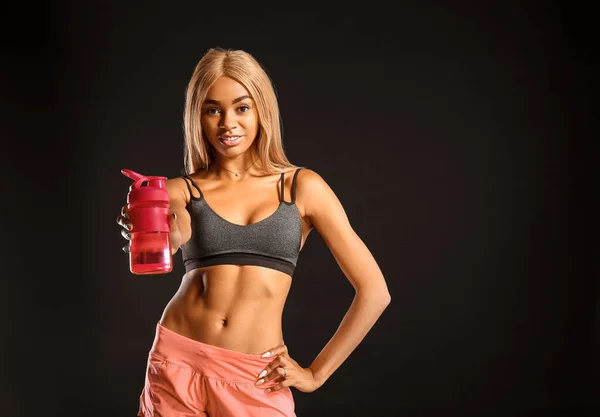 Sporty young African-American woman with bottle of water on dark background — Stock Photo, Image