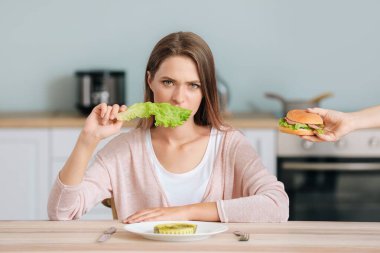 Üzgün kadın sağlıklı marul yiyor ve kadın eli ona lezzetli bir burger sunuyor. Diyet kavramı