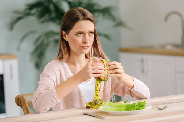 Mulher estressada segurando hambúrguer não saudável com fita métrica na cozinha. Conceito de dieta — Fotografia de Stock