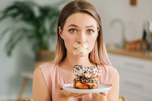Stressed woman with taped mouth and with tasty donuts in kitchen. Diet concept — Stock Photo, Image